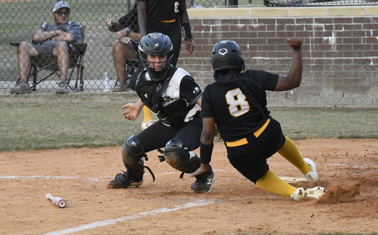 A softball catcher prepares to tag a sliding runner at home plate during a game, with spectators watching from behind a fence.