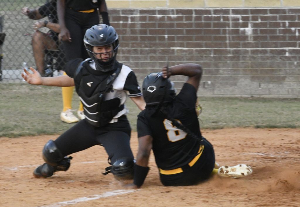 A catcher in protective gear attempts to tag out a sliding softball player wearing a helmet and black uniform at home plate during a game.