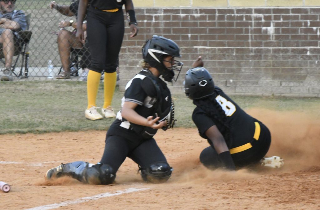 A softball catcher tags a sliding runner at home plate, kicking up dust, as another player and spectators watch in the background.