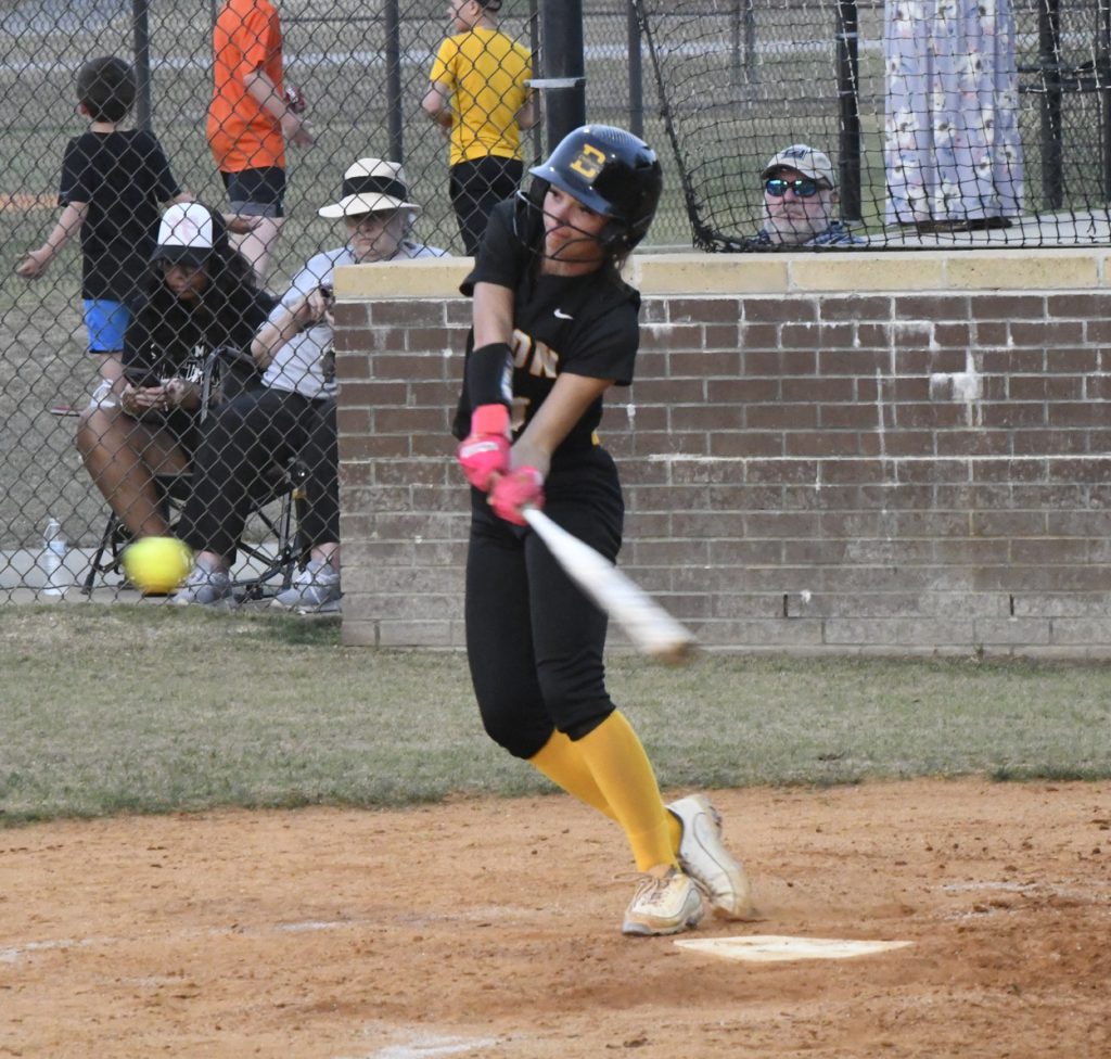 A youth softball player in black and yellow uniform hits a pitched ball, with spectators watching behind a fence.