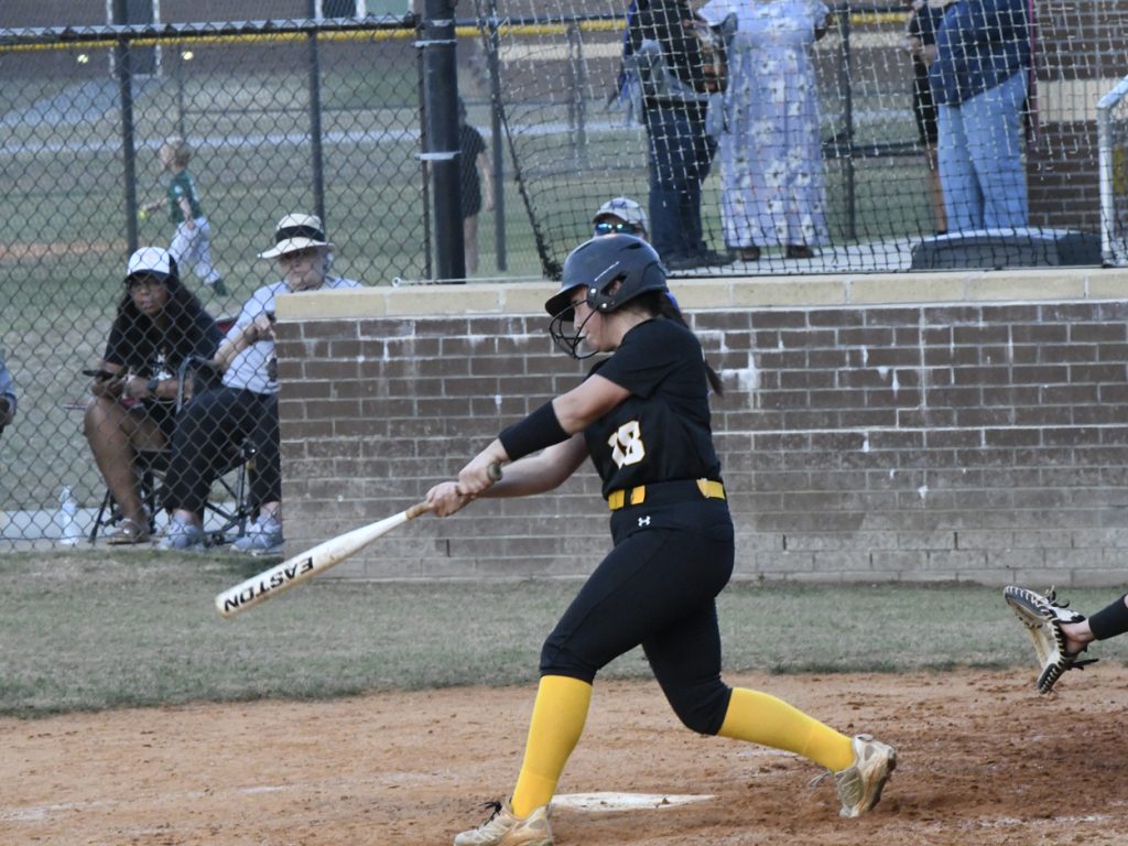 Softball player in black and yellow uniform swings bat during a game, with spectators seated behind a chain-link fence in the background.