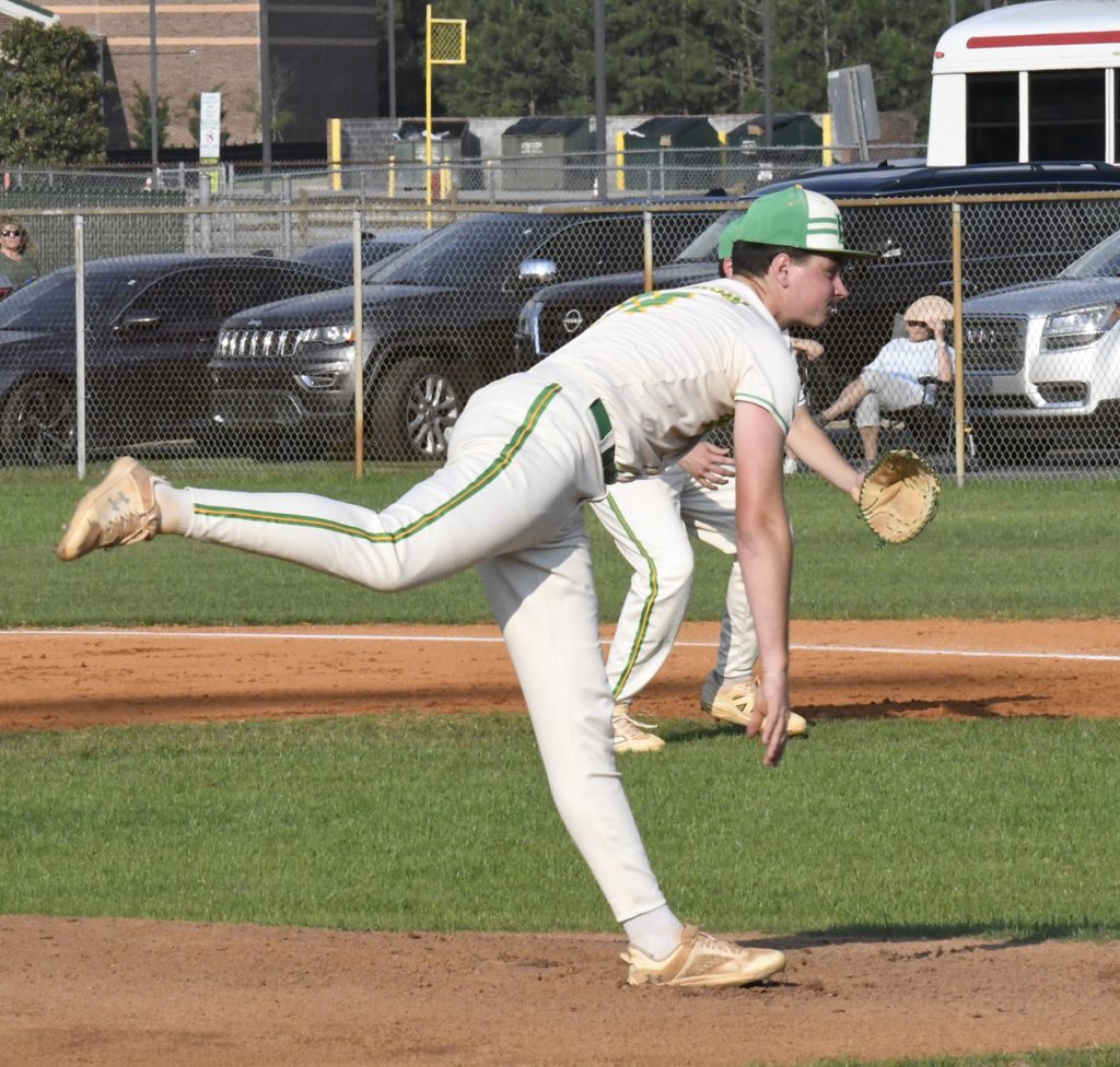 A baseball pitcher in white and green uniform throws a pitch during a game on a sunny day, with cars parked in the background.