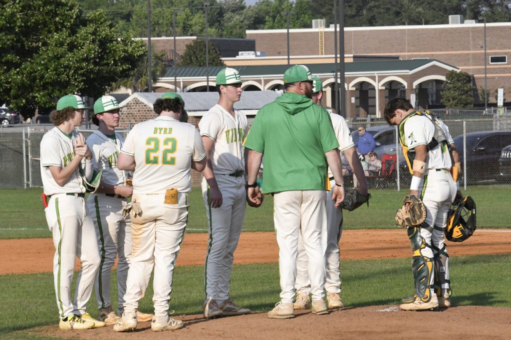 A group of baseball players and a coach huddle on the pitcher's mound during a daytime game, with school buildings visible in the background.
