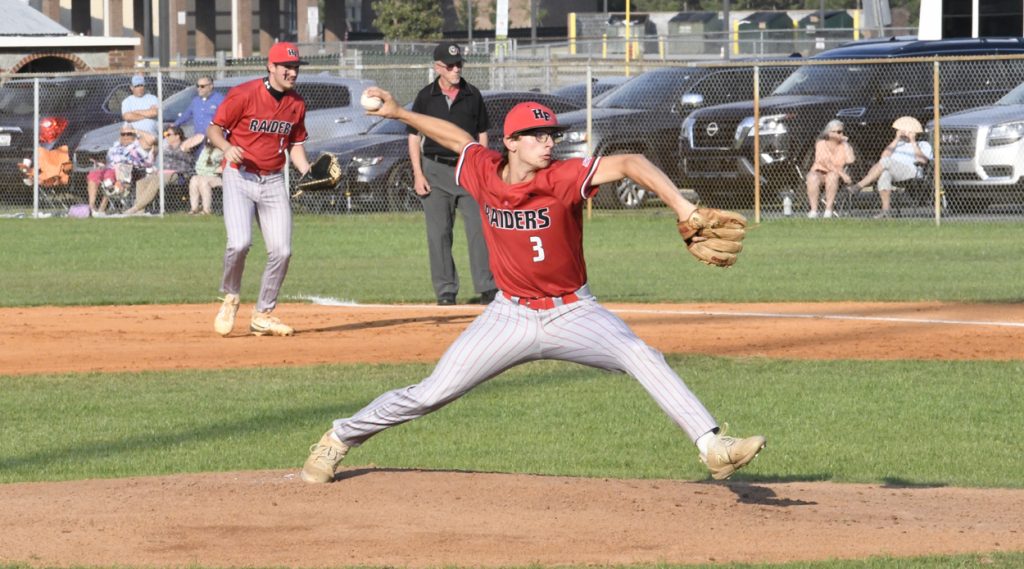A baseball pitcher wearing a red "Raiders" uniform throws a pitch on a sunny day, with teammates and spectators in the background.