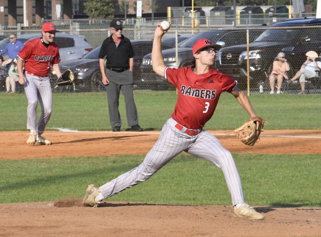 Baseball player in red uniform pitching the ball, with teammate, umpire, and spectators in the background on a sunny day.