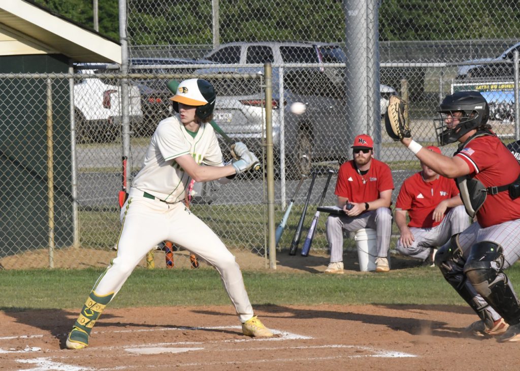 A baseball batter prepares to swing at a pitch, with a catcher and coaches in red uniforms watching from behind home plate.