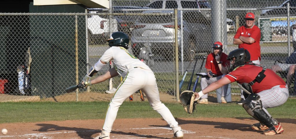 A baseball player in white bunts the ball while the catcher in red prepares to catch, with coaches and players watching from the dugout and stands in the background.