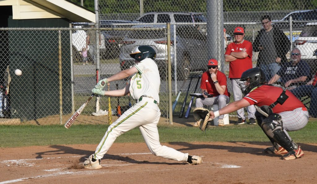 A baseball player in white swings at a pitch while the catcher and umpire watch closely, with spectators and coaches in the background.