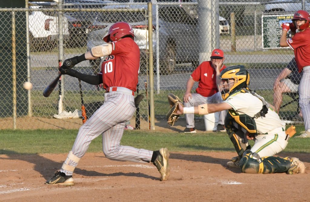 Baseball player in a red uniform swings at a pitch, while the catcher in green and yellow prepares to catch the ball during a game.