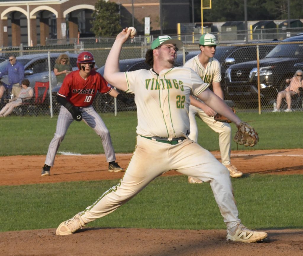 A high school baseball pitcher in a white "Vikings" uniform throws a pitch, with a runner leading off first and teammates in the background.