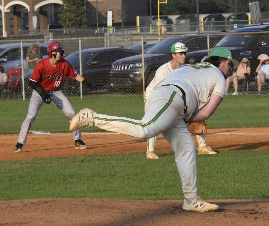 A baseball pitcher in white throws a pitch as a baserunner in red prepares to run, with teammates and spectators in the background.