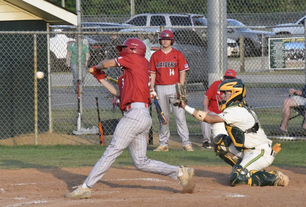 A baseball player in a red uniform swings at a pitch, while the catcher in yellow and green gear is ready behind home plate.