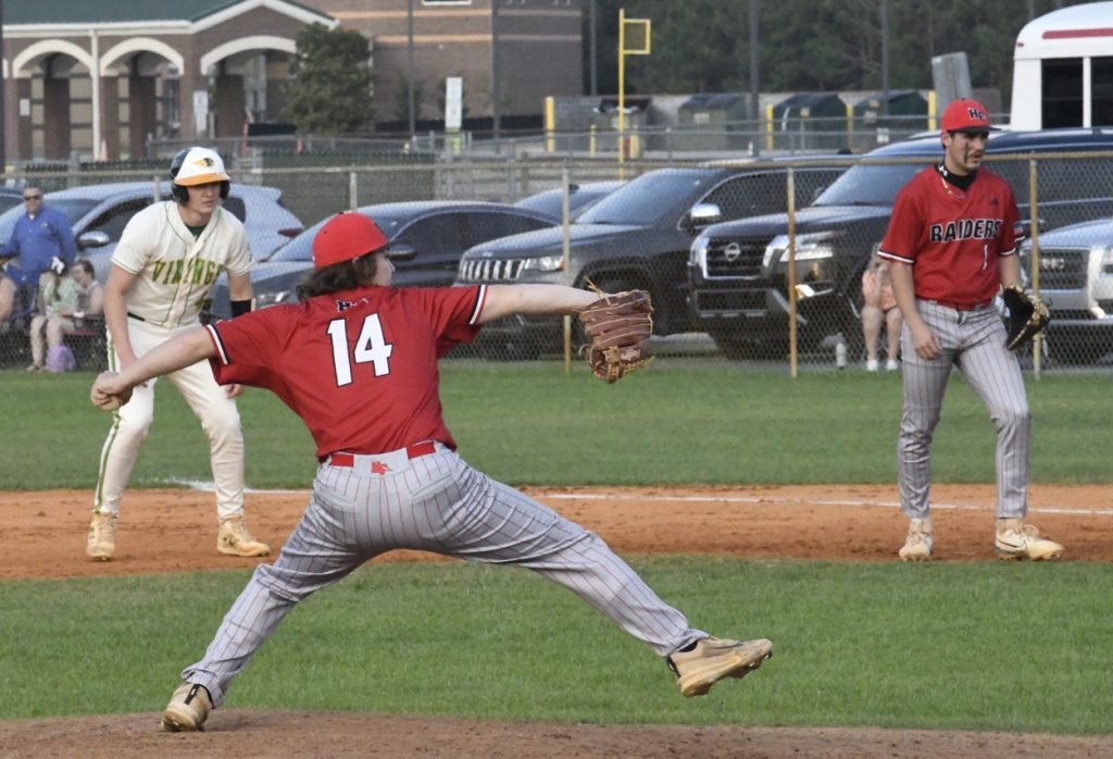 A baseball pitcher in a red uniform throws the ball while a runner and fielder watch, with cars and people in the background.