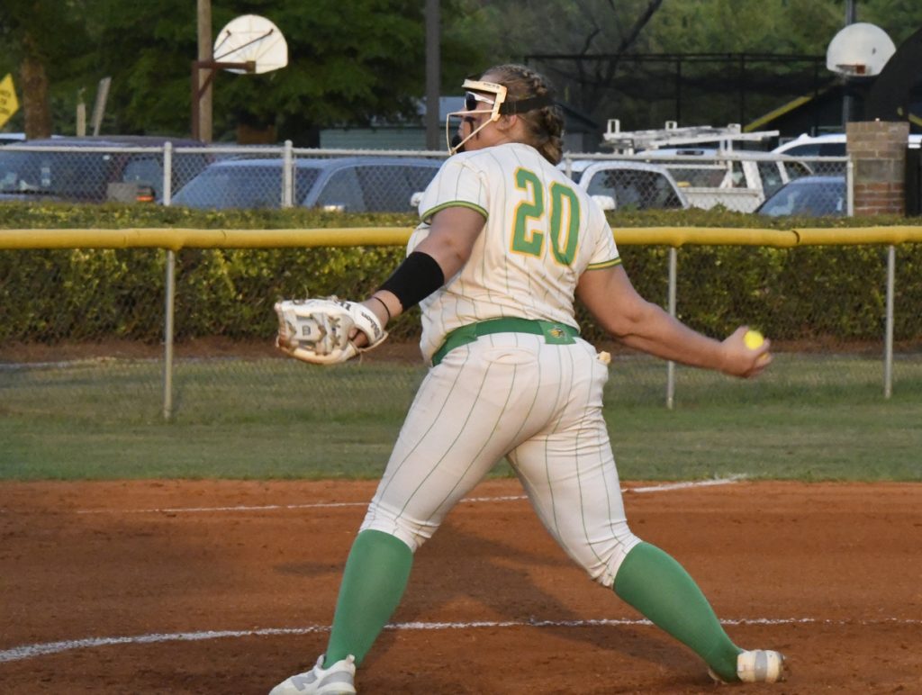 A softball player wearing number 20 winds up to pitch on a dirt field, with green socks and white uniform, during a game.