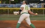 A softball player wearing number 20 winds up to pitch on a dirt field, with green socks and white uniform, during a game.