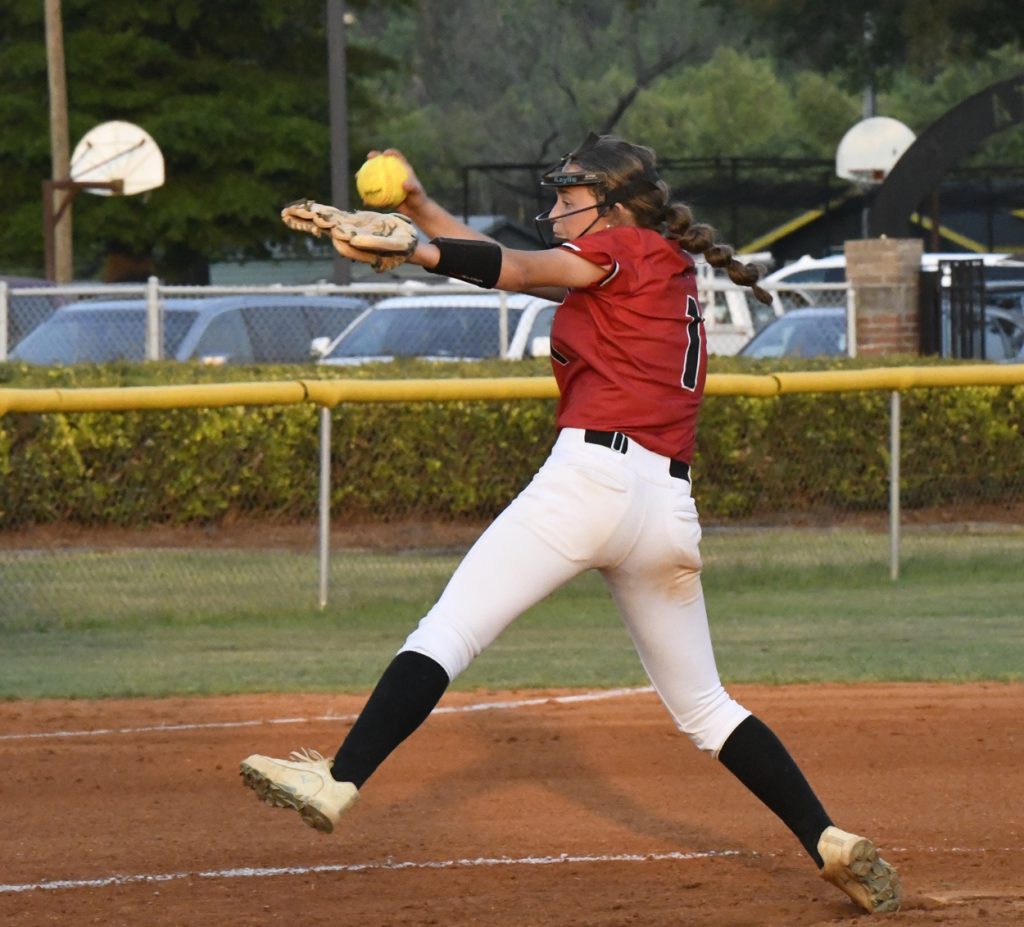 A softball pitcher in a red jersey and white pants winds up to throw the ball on an outdoor field.