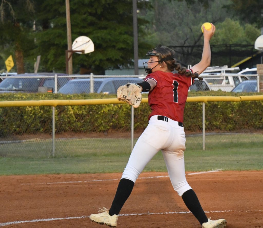 A softball pitcher wearing a red jersey and white pants winds up to throw a pitch on an outdoor field.