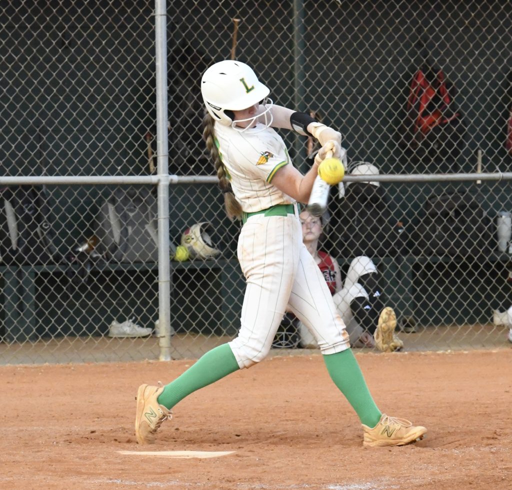 A softball player in white and green uniform hits a yellow softball with a bat, action taking place on a dirt field near a chain-link fence dugout.