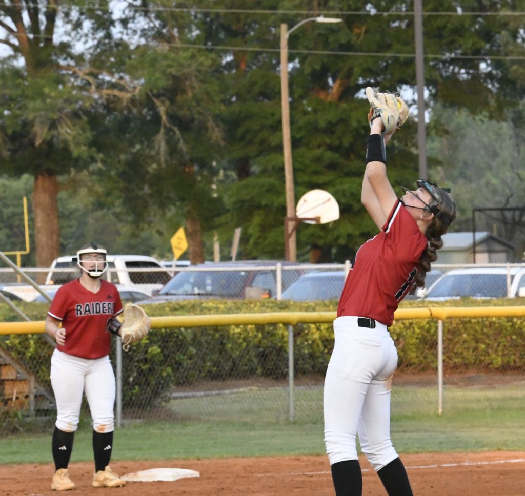 Two softball players in red jerseys; one is about to catch a softball, the other watches nearby on a dirt field with cars and trees in the background.