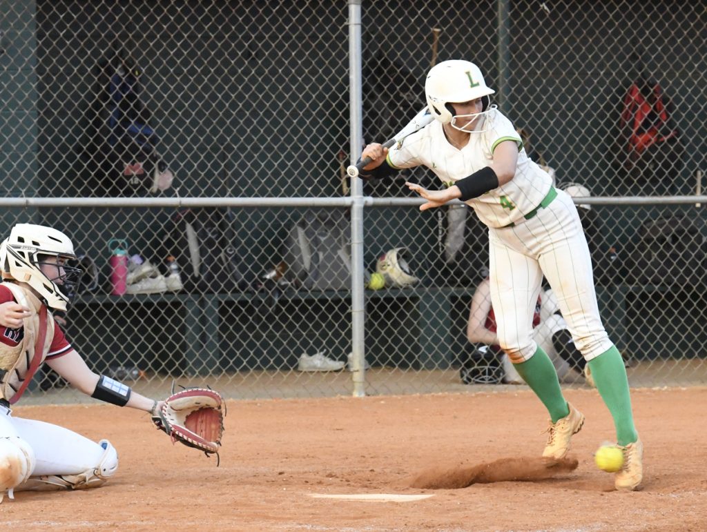 Softball player bunts the ball as the catcher prepares to field it during a game, with a dugout and fence in the background.