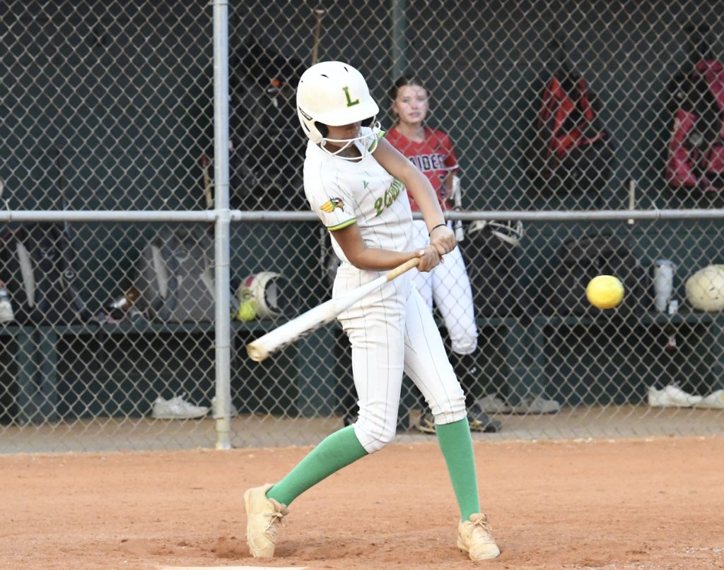 A softball player in white uniform and helmet hits a yellow softball during a game, with teammates and equipment in the dugout behind a chain-link fence.