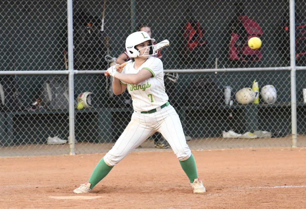 Softball player in white and green uniform prepares to swing at an incoming pitch on a dirt field, with dugout equipment visible in the background.