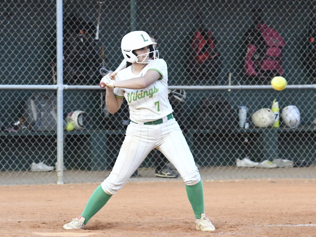 Softball player in white and green uniform prepares to hit a pitched ball during a game, with dugout and gear visible in the background.