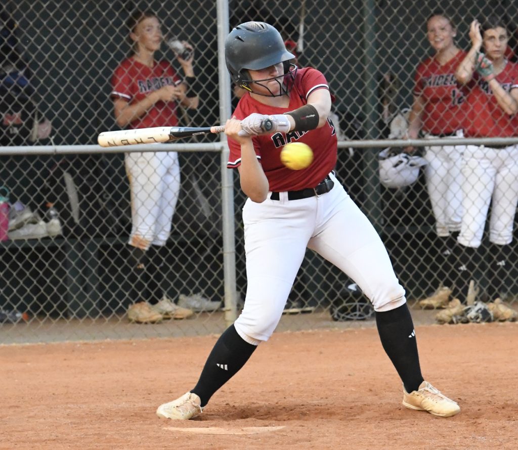 Softball player in red jersey and white pants swings at a yellow ball. Teammates in the dugout watch through a chain-link fence.