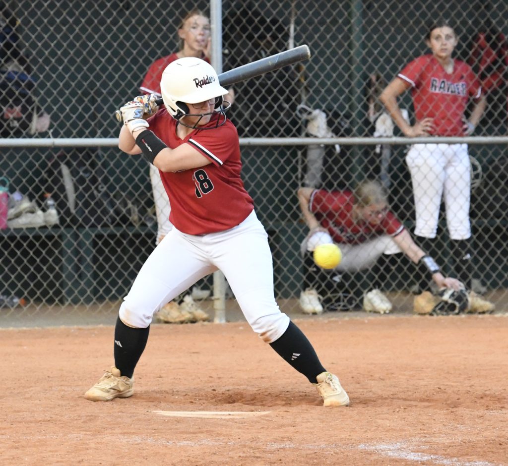A softball player in a red jersey prepares to bat, while teammates watch and one retrieves a ball in the dugout.