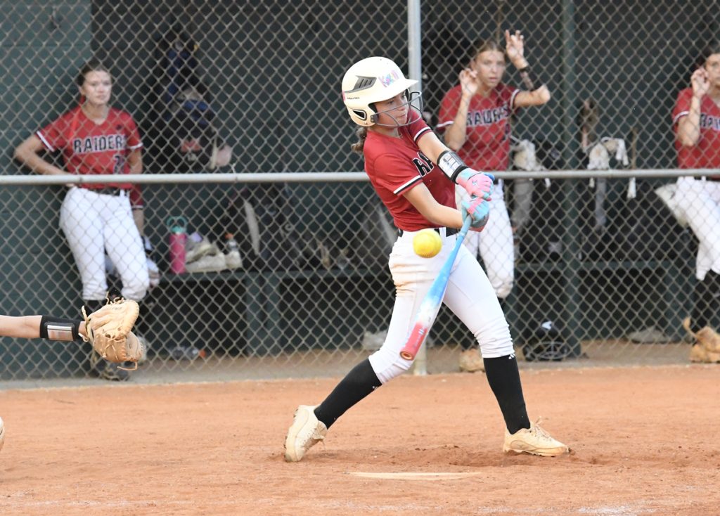 A softball player in a red jersey swings at a yellow ball during a game, with teammates watching from the dugout behind a chain-link fence.