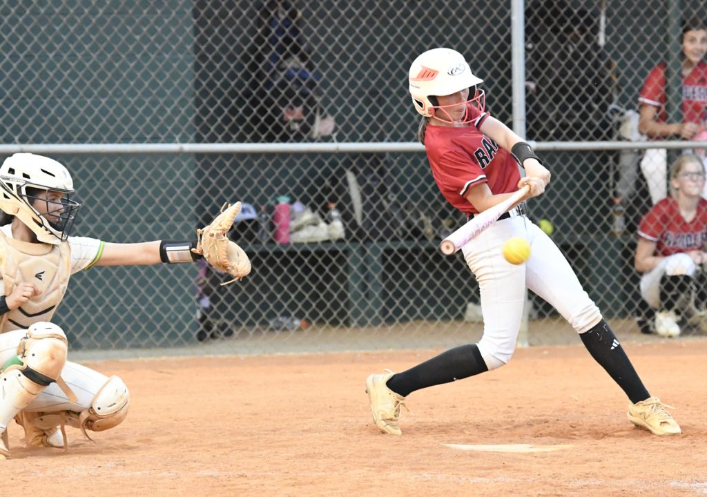A softball player in a red jersey swings at a yellow ball while a catcher in protective gear reaches to catch it.