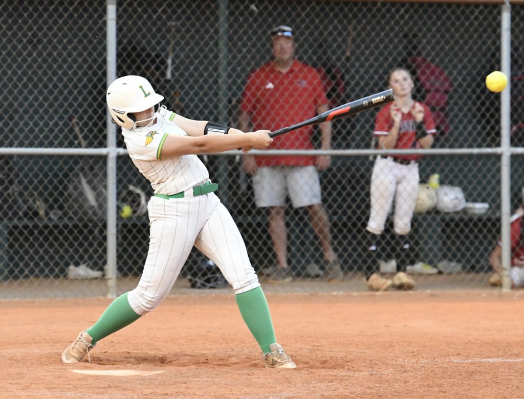 Softball player in white uniform swinging a bat at a yellow ball during a game, with teammates and coach watching from behind a chain-link fence.