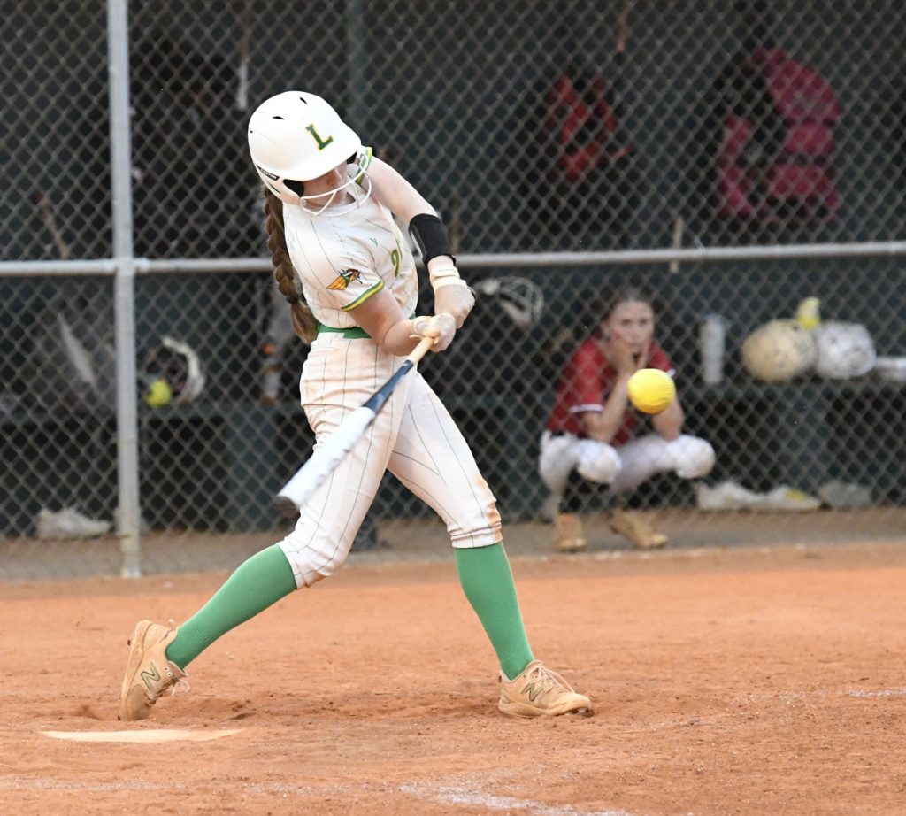 A softball player in a white and green uniform swings at a yellow ball, with a catcher and equipment visible in the background.