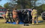 Firefighters in full gear respond to a car accident, standing near an overturned vehicle on its side with equipment on the ground.