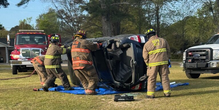 Firefighters in full gear respond to a car accident, standing near an overturned vehicle on its side with equipment on the ground.