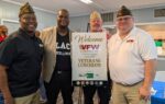 Four men, some in VFW hats, stand smiling by a sign welcoming guests to a Veterans Luncheon event at a VFW hall.