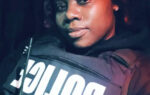 A person wearing a police vest and holding a radio, looking at the camera with a neutral expression in low light.
