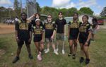 A group of six young adults pose outdoors on a grassy field, some wearing matching black and gold outfits, with trees and a fence in the background.