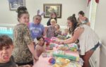 A group of children and adults gather around a table with party supplies, smiling and celebrating at an indoor event with decorations.