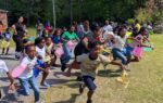 A group of children excitedly running outdoors during an egg hunt event, holding colorful baskets, with adults and trees in the background.