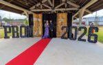 A couple stands on a red carpet under a pavilion decorated with large "PROM 2026" letters for a prom event.
