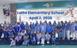 A group of adults posing together inside a school gym, under a sign reading “Latta Elementary School, April 2, 2026.”