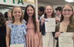 Four smiling girls in dresses hold up certificates in a classroom or school setting, with other students and decorations visible in the background.