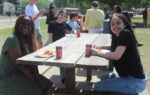Three women sit at a picnic table outdoors, smiling, with food and drinks. Other people are gathered in the background on a sunny day.