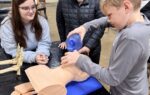 A boy practices a medical procedure on a mannequin while three other students watch attentively at a classroom table.