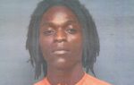 A person with shoulder-length dreadlocks wearing an orange shirt poses in front of a plain, blue-gray background.