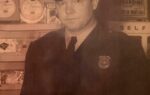 A police officer in uniform stands indoors in front of shelves with various items, in a sepia-toned vintage photograph.