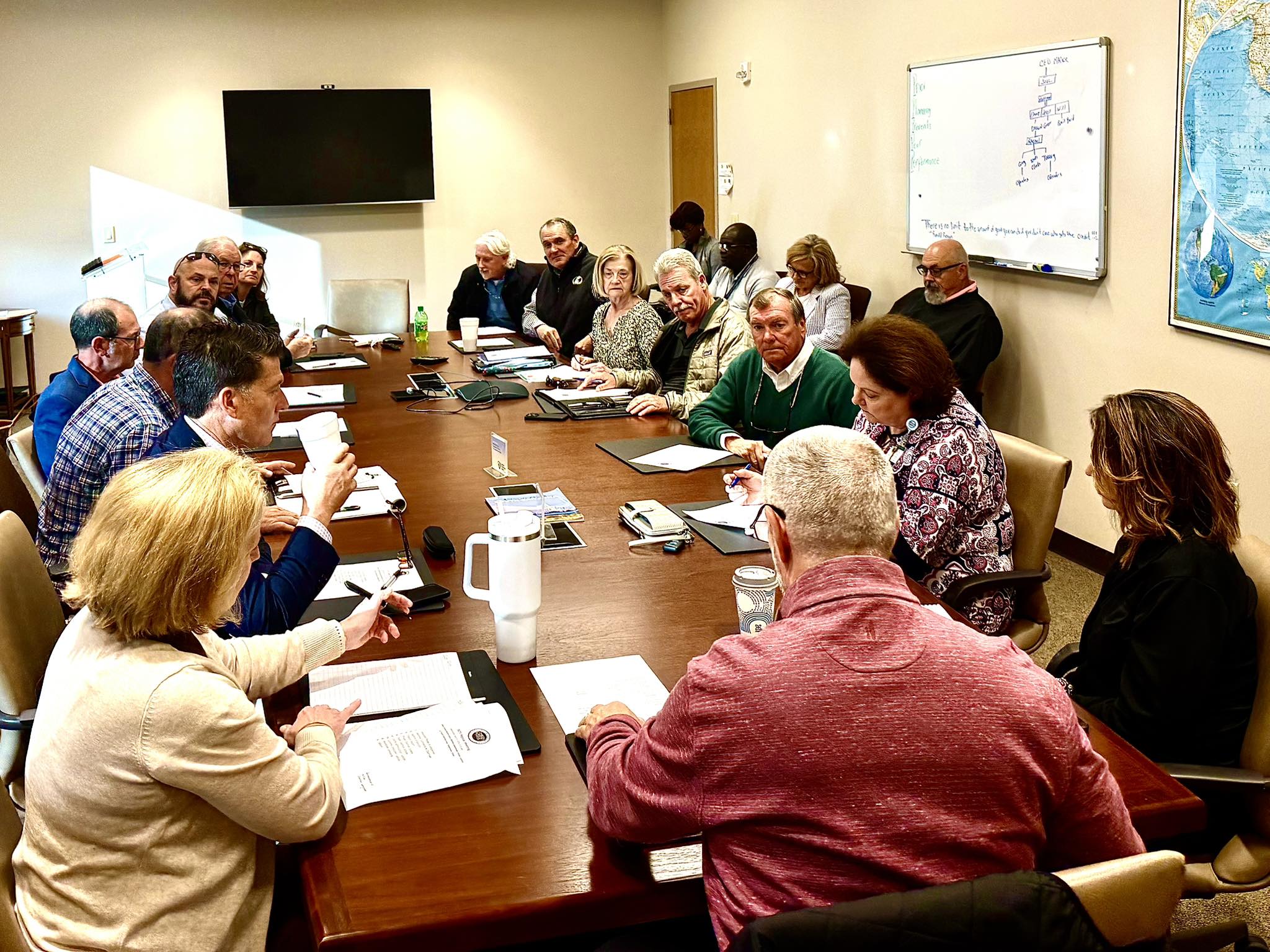 People sitting around a conference table having a meeting, with papers, pens, and coffee mugs on the table, and a large monitor on the wall.