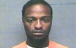A man with braided hair wearing an orange prison uniform poses for a mugshot against a gray background.