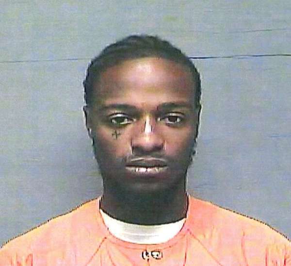 A man with braided hair wearing an orange prison uniform poses for a mugshot against a gray background.